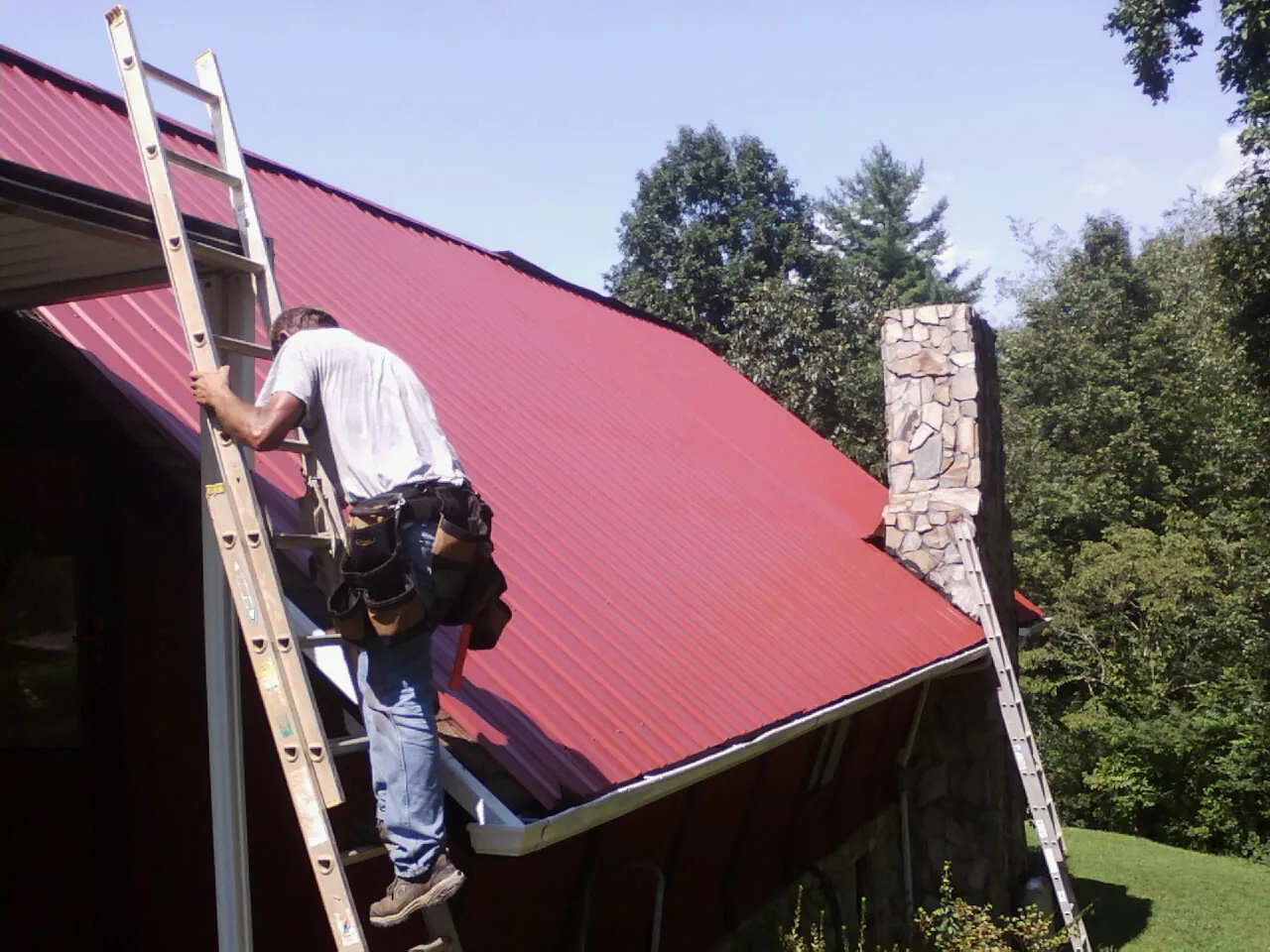 Roofer on a ladder beside a red metal roof and stone chimney — Soffit & Fascia Repair by Roofing Brothers in Bradfordwoods